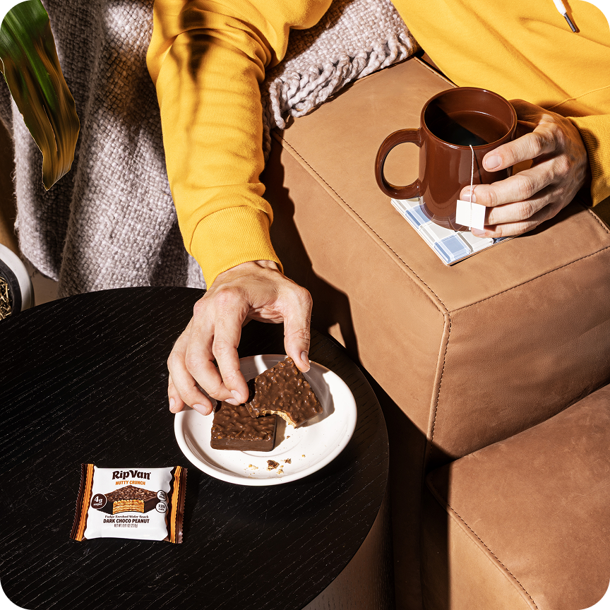 Person holding a broken dark chocolate wafer bar on a white plate with a packaged bar and a cup of tea